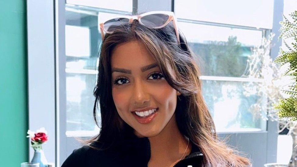 A head and shoulders photo of Sanam Harrinanan smiling for the camera in a cafe. She has a pair of sunglasses on the top of her head and her brunette hair is worn down. She is wearing a black top and behind her is a window. You can just make out a small vase of flowers on another table and a fern plant to the right-hand side of her head