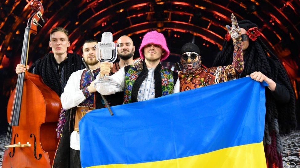 The members of Kalush Orchestra standing on a stage, celebrating while holding the Ukrainian flag