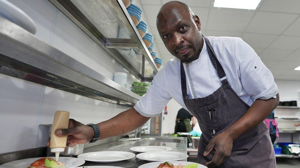 Chef George Opondo looks at the camera as he applies garnishes to dinner plates in a large kitchen