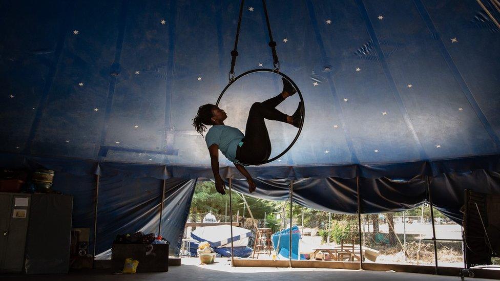 Female acrobat in a balancing herself in a suspended wheel. She is in a circus tent.