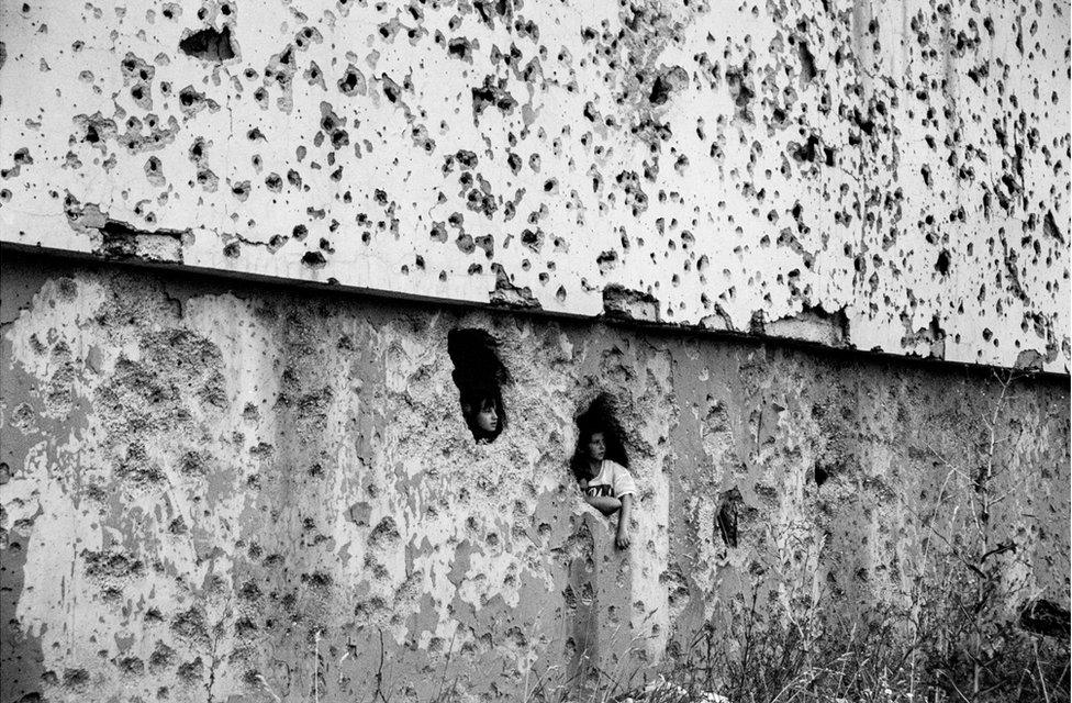 Children look out of a holes in a wall that is covered in bullet holes