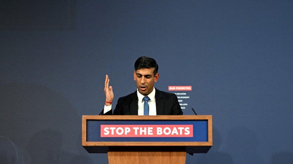 Prime Minister Rishi Sunak stands above a "stop the boats" sign