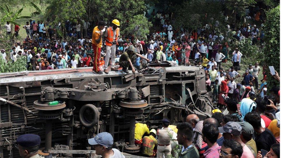 Dozens of local people gathering near an overturned train carriage, with three emergency service workers standing on top of it