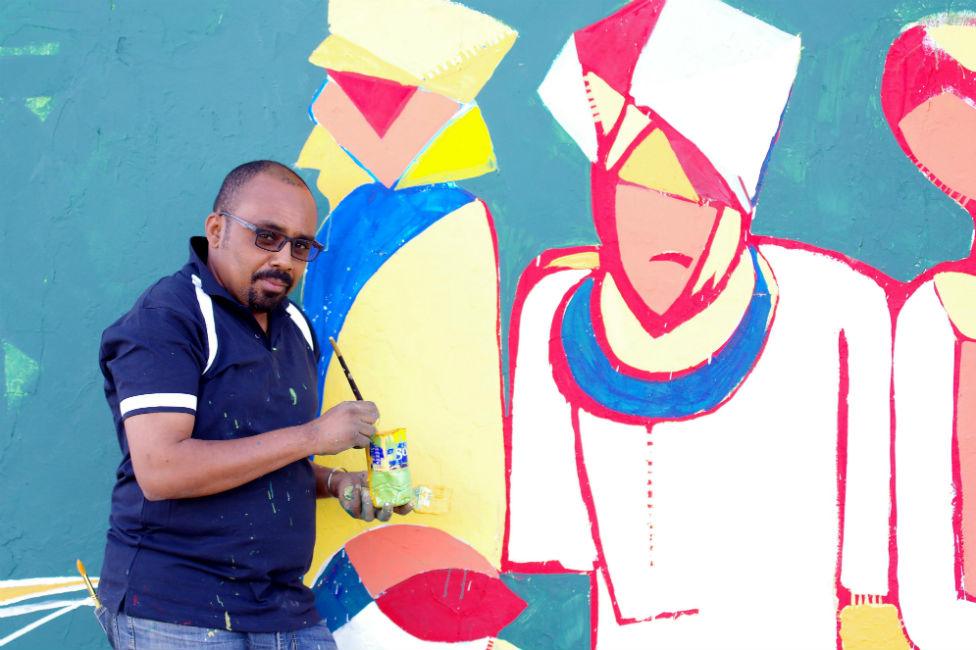 A man stands in front of a mural with paint and a brush in his hands