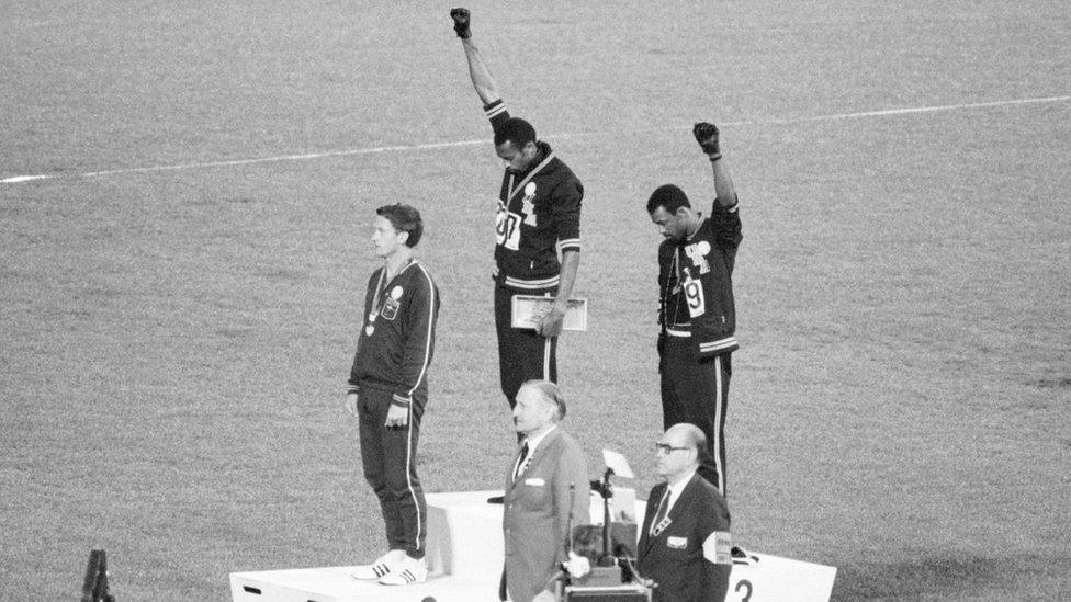 Athletes Tommie Smith and John Carlos - stand with their heads bowed and eyes closed, their hands raised with black gloves, and fists clenched.