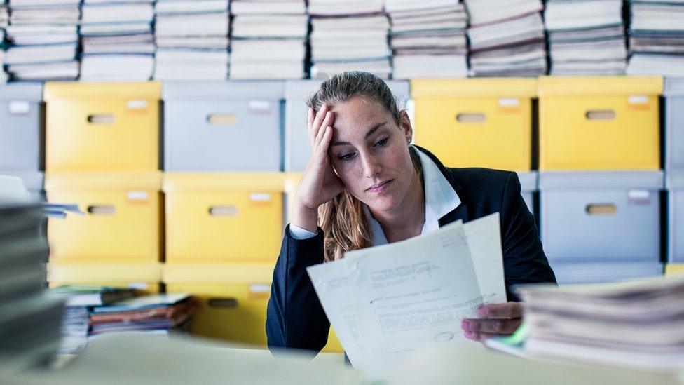 A woman reads documents in a cluttered office