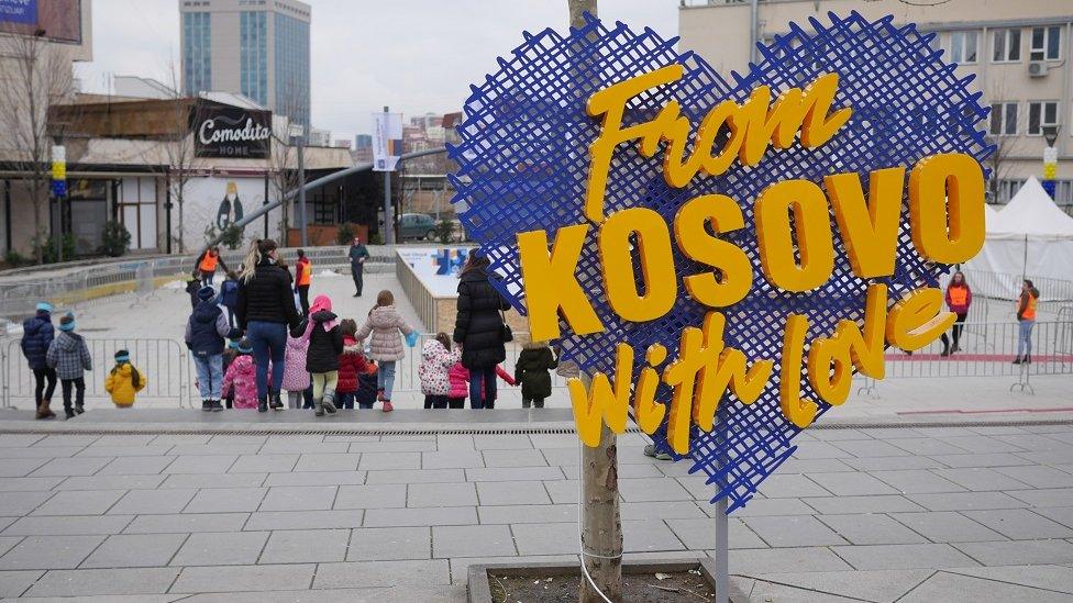 A sign saying 'From Kosovo with Love' stands on a street with children in the background.