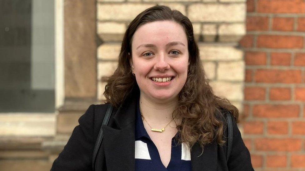 Nichole Brown. Nichole is a 25-year-old white woman with long curly brown hair. She has blue eyes and smiles at the camera. She wears a black coat over a blue and white striped top with a gold necklace and a black back pack on her shoulders. She's pictured outside in Stoke-on-Trent in front of a brick wall