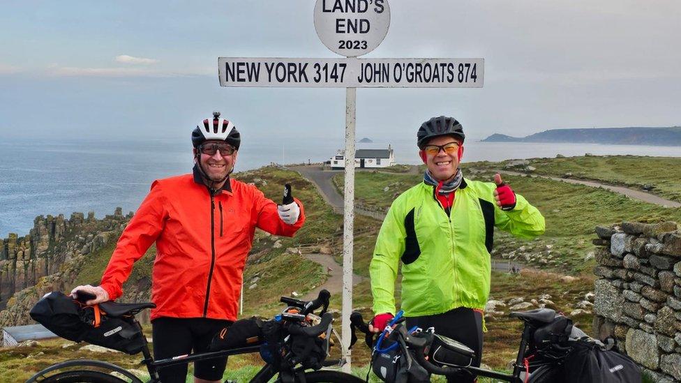 Mark and Ian standing with their bikes in front of the landmark sign at Land's End, Cornwall.