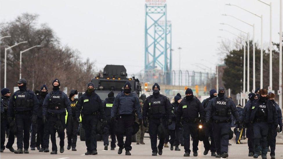 A row of black-clad police in riot gear walk down a road towards the protesters' encampment