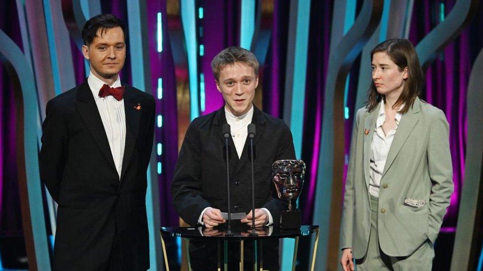 Young man with light-coloured hair flanked by man and woman at awards ceremony