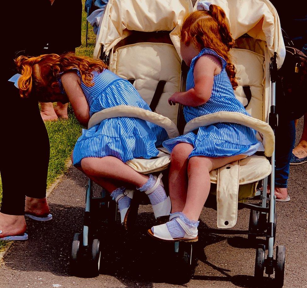 Twin girls in matching blue dresses lean over to retrieve their snacks at the bottom of the buggy