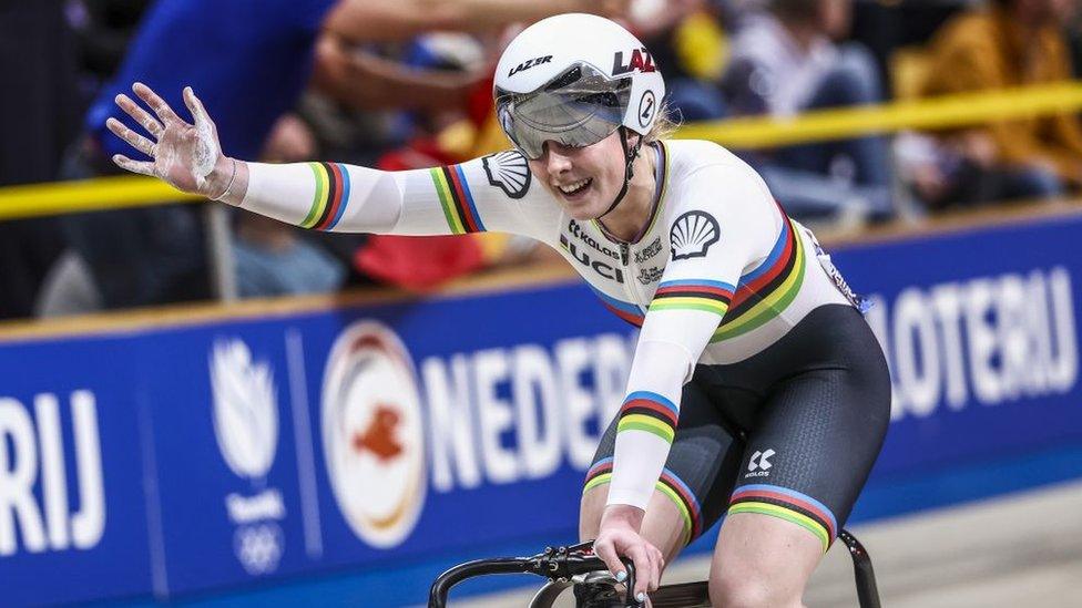 Britain's Emma Finucane competes in the Women's Sprint qualifying round race during the second day of the UEC European Track Cycling Championships at the Omnisport indoor arena in Apeldoorn, on January 11, 2024. Emma wears a white cycling jersey with green, yellow, black, red and blue stripes on her arms and wrists. She wears a white helmet with a black visor. She holds her left arm out towards spectators, her left hand on the bike's handlebars. Emma is a 21-year-old white woman and is pictured smiling.