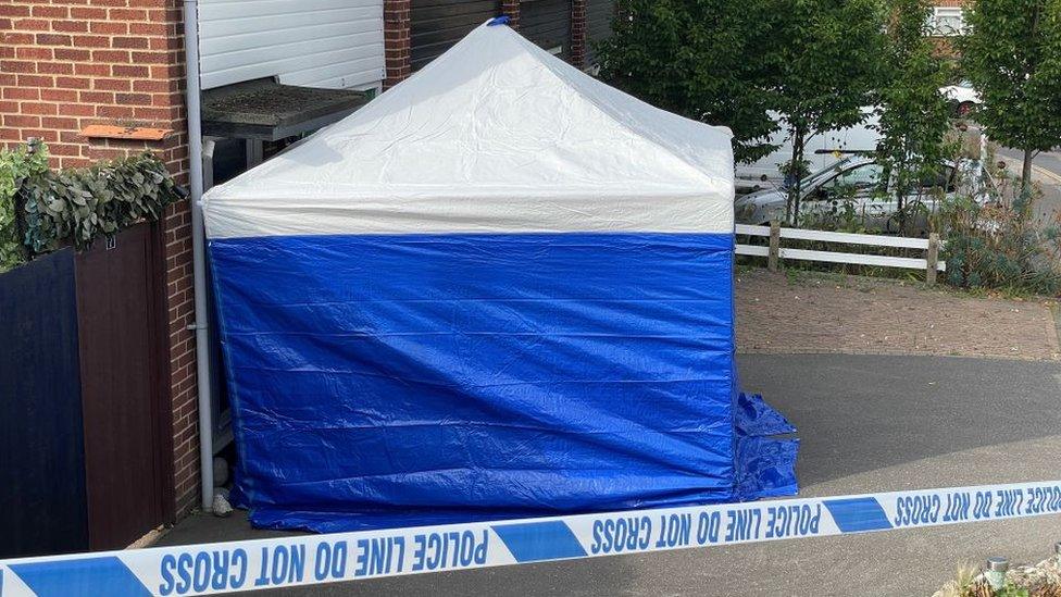 A white and blue police tent is outside a house, behind police tape.