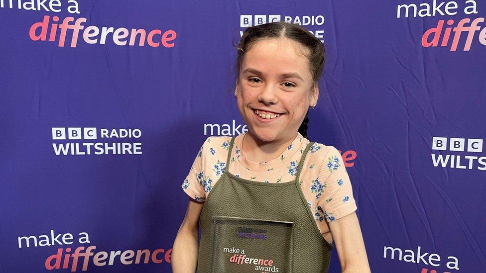 A little girl in front of a purple banner holding a glass award.