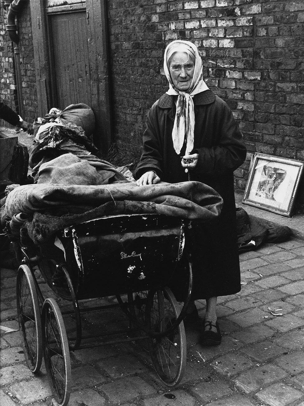 An elderly woman stands next to a pram with bundles of cloth on