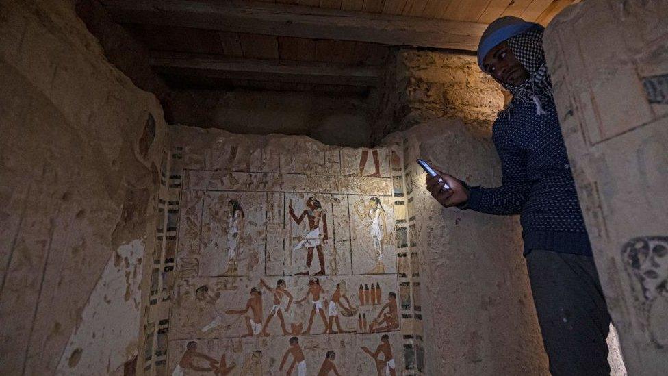 man shining phone torch on ancient Egyptian painting and carving in wall of tomb.