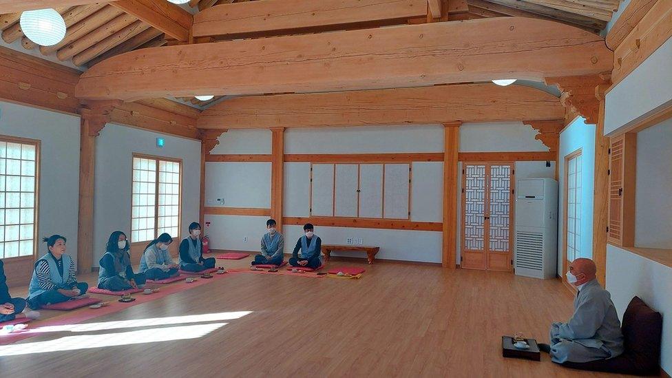 A group of men and women sit cross-legged facing a monk in a sparsely-decorated room in a Buddhist temple