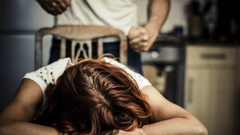 A man stands with a clenched fist above a woman who lies her head on the table.