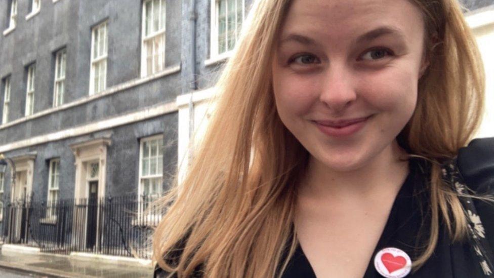 A young woman with long blonde hair stands in front of 10 Downing Street - a long street with, flat dark grey frontage and large windows with white painted frames. She's smiling, and wearing a smart black jacket with a round badge showing a red heart.