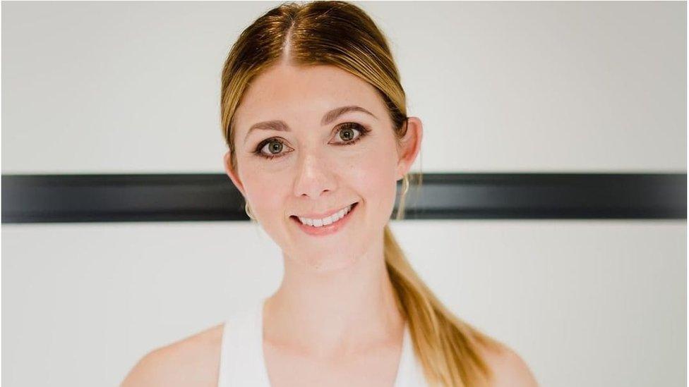 Nikki Franklin, smiling, stands against a mostly white background with a thick black band passing horizontally through the middle. Her hair is tied back in a long ponytail, and she's wearing a white vest top.