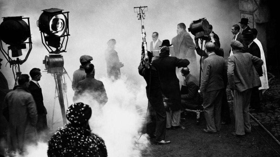 Black and white photo of filming, with dense fog, lighting and actors