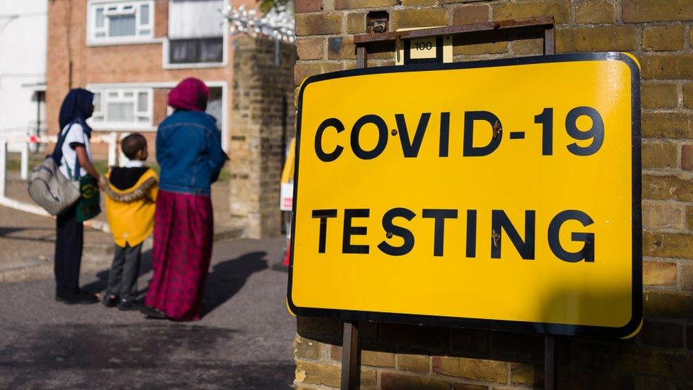 A sign saying COVID-10 TESTING with a family of three queuing beside it