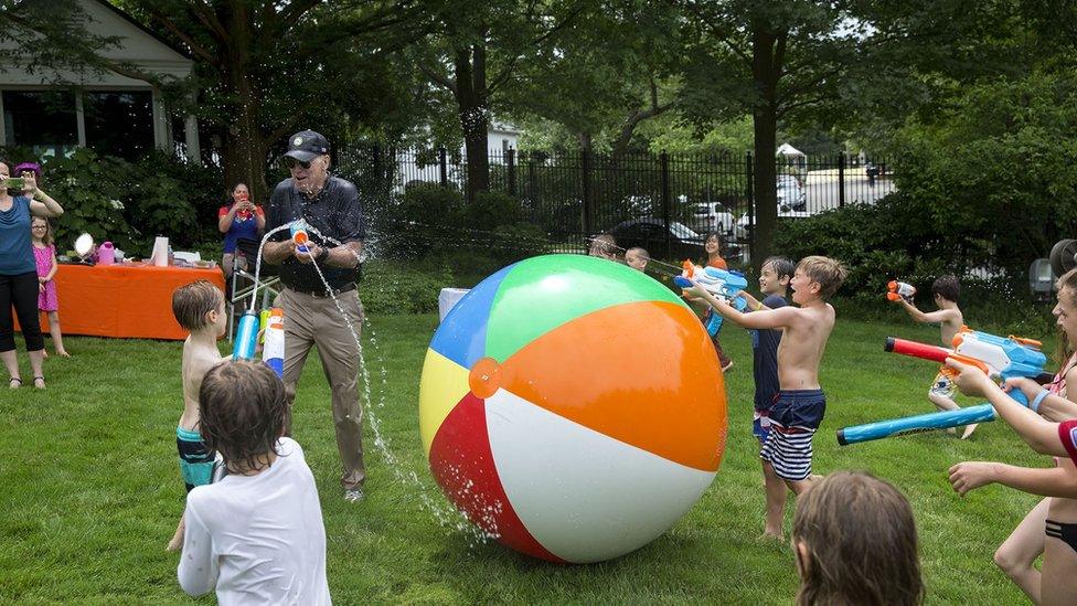 A ring of children spray vice president Joe Biden with a water gun, which he returns in kind. A giant, chest-high beach ball lies between them.