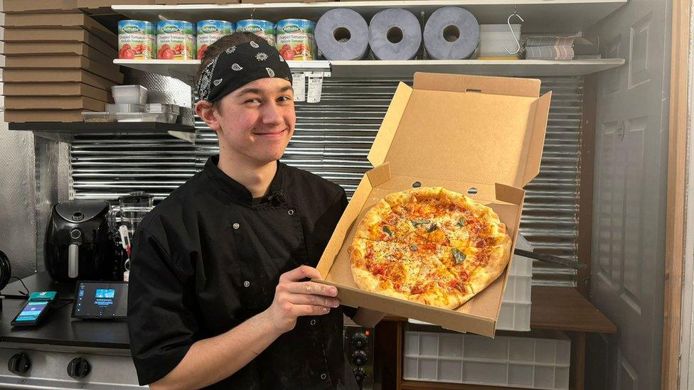 Joshua holding up a pizza and smiling at the camera. He is wearing a black button up chefs jacket and a bandana