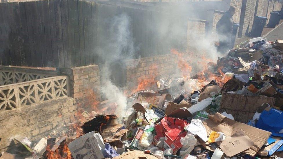 The bin wagon's rubbish dumped in a back lane with flames and smoke rising