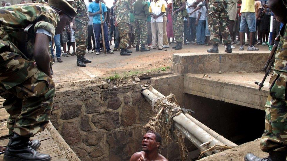 A bloodied man in a pit begs as he is surrounded by soldiers.