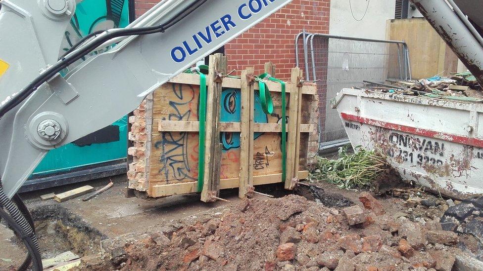 The section of wall, boarded up and surrounded by a crane and skip