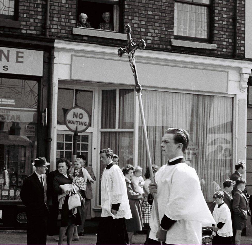 Residents, including two women from an upstairs window, watch a church procession