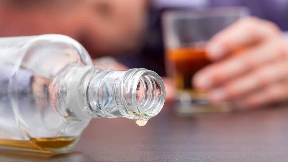 An empty bottle of alcohol on its side in the foreground - a man holding a glass of alcohol in the background