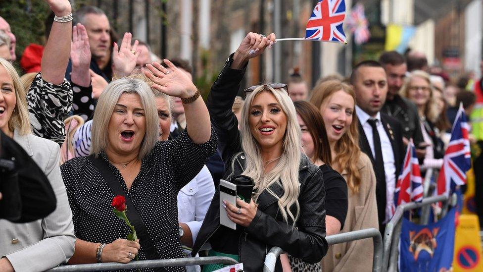 A crowd of people - some waving, holding flags and cheering - gathered outside Hillsborough Castle ahead of the visit by the King and Camilla