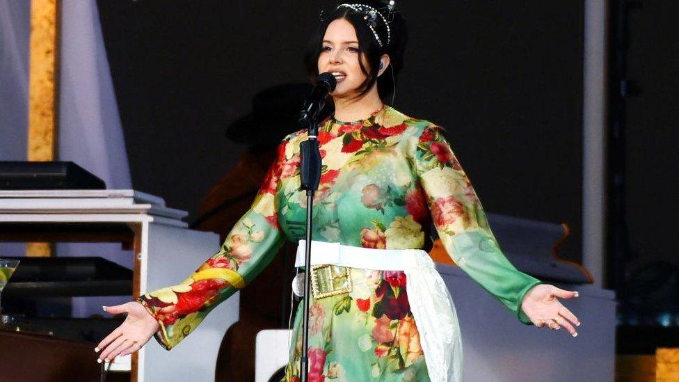 Lana Del Rey singing on stage, she is wearing a green floaty dress and her arms are open. She is wearing her black hair up.