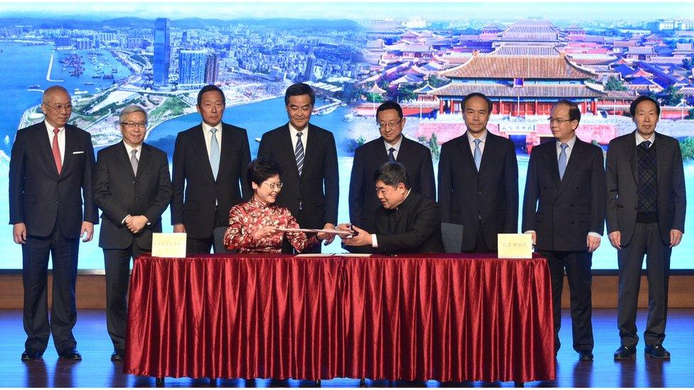 Ms Lam seated at the signing ceremony, with one man seated next to her and eight officials standing behind.