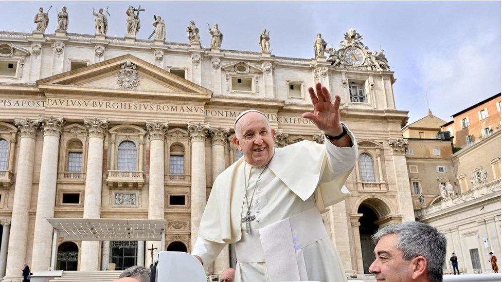 Pope Francis with hand up in front of Vatican building