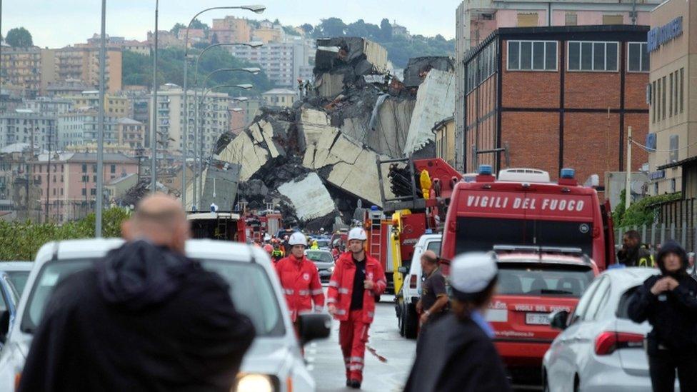 Large mound of debris is shown blocking a road, surrounded by emergency services