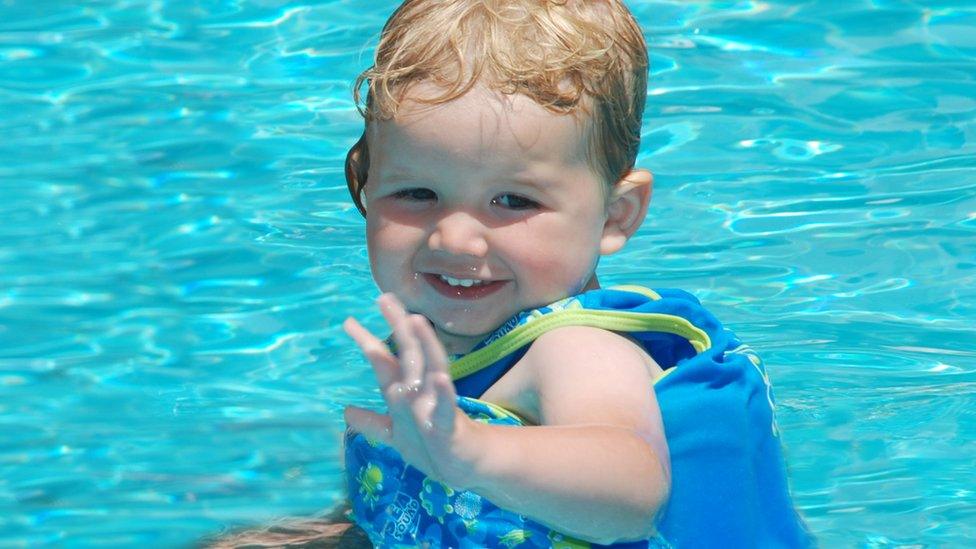 A joyful picture of a toddler wearing a blue floatation device and the swimming pool. He's waving at the camera.