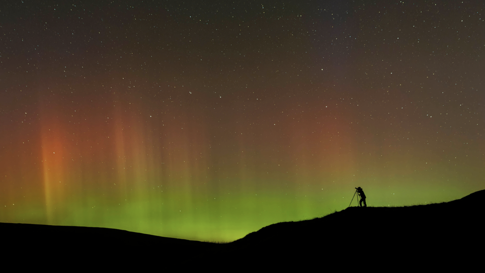 Silhouette of a person on a hill with camera equipment looking up into the night sky where there are the green and red hues of the aurora.