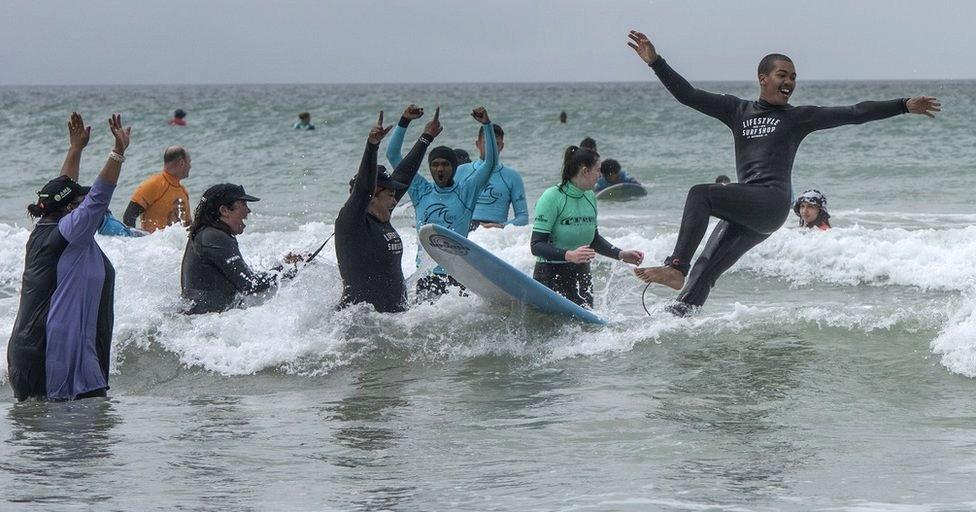 A young man smiles and stretches out his arms as he falls off a surfboard into the sea. He is surrounded by smiling adults.