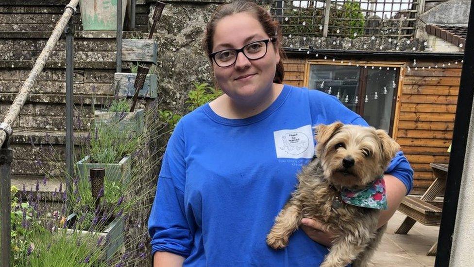 A white young woman wearing glasses holding a dog under her arm in a garden