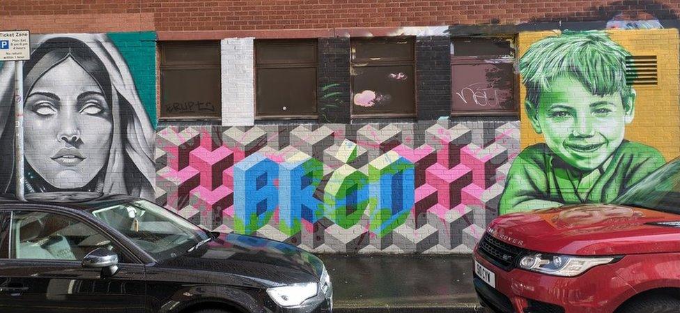 Paintings of two people and the word Bród on a wall behind two parked cars