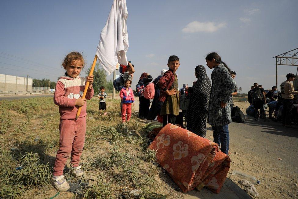 A young girl holds an improvised white flag as her family, heading south, stops to rest on the Salah al-Din road.