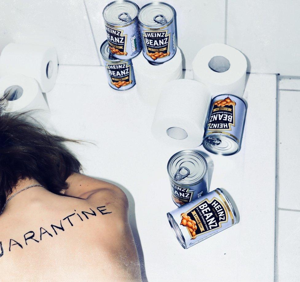 A woman lies in a bath surrounded by cans of baked beans with the word quarantine on her back