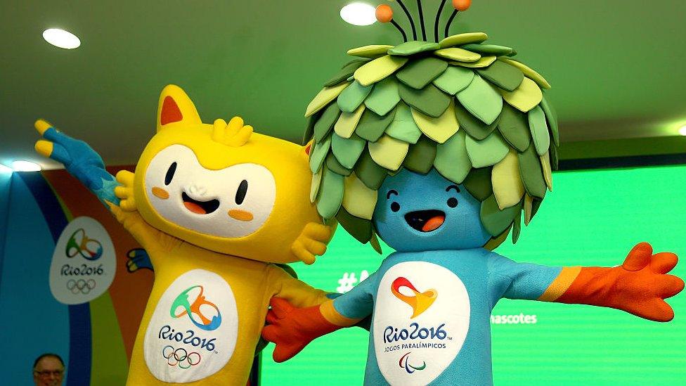 Vinicius is a yellow cat-like mascot with blue hands. Tom has what looks like a shrub on his head and antenna coming out the top. He has blue skin and looks happy