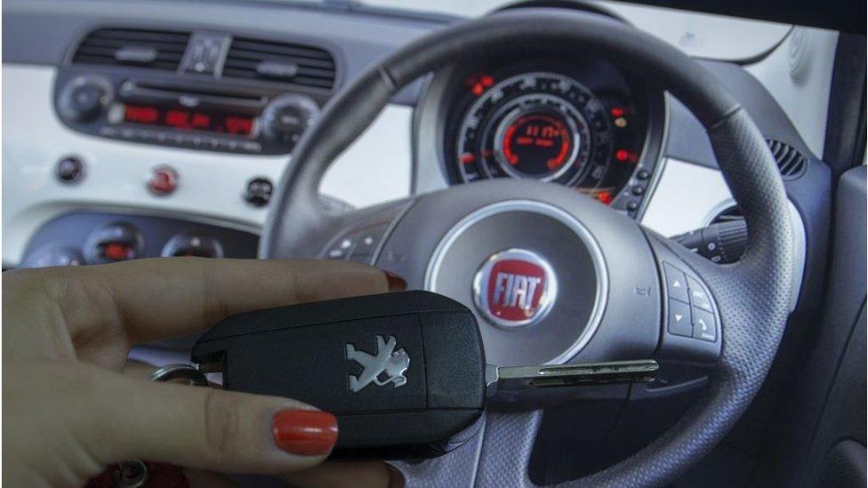 A woman holds a Peugeot car key in front of a Fiat steering wheel