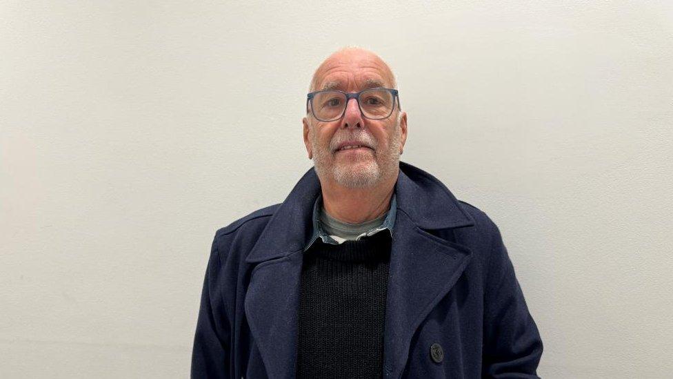 Man with very short hair and short grey beard wearing a blue raincoat