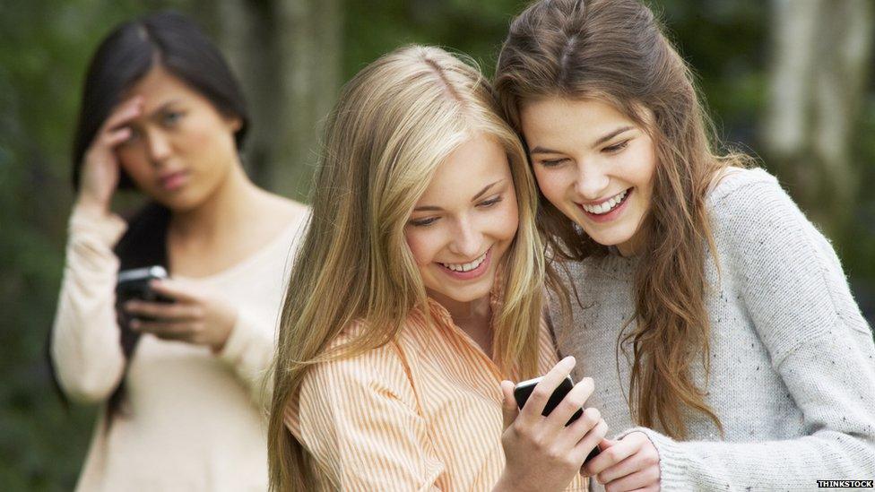 Two girls smiling and looking at a phone while another looks sad